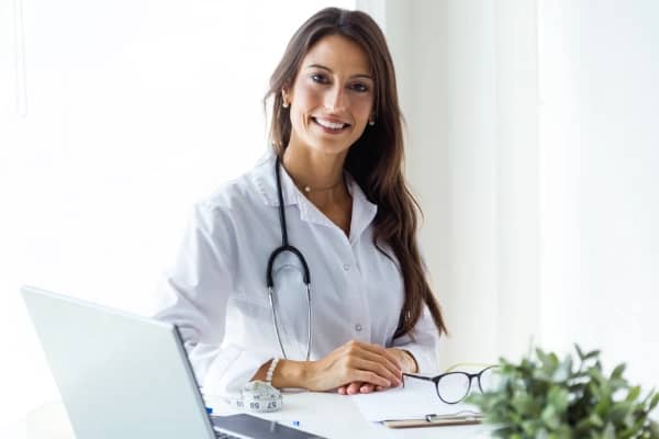 A smiling primary care female doctor sitting at her desk looking at the camera